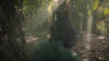 Siyah elbiseli genç sıcak sarışın kadın poz ve palmiye ağaçları ve yeşil yaprakları yakınında kalın duman dans. Yere düşen güneş ışığı. Bali 