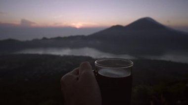 Adam el yanardağ, dağlar ve gündoğumu arka plan sıcak kahve ile cam tutar. Sabahın erken saatlerinde silüeti ve gölün tepesinden güzel bir manzara. Bali, Endonezya