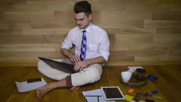 年轻的严肃的男人在家里工作 在木墙的背景滑稽的裤子 赤脚的家伙坐在地板上 看着笔记本电脑和打字 — 图库视频影像