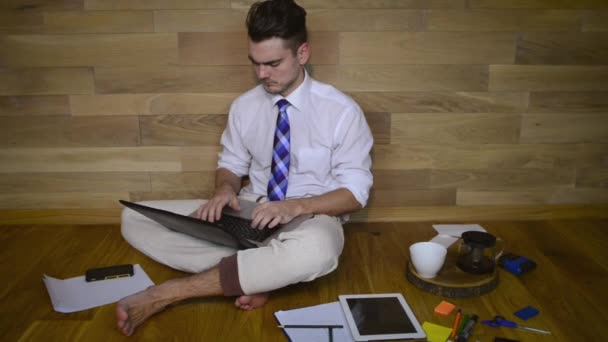年轻的赤脚男子在家里工作 穿着睡衣裤 坐在地板上的木墙背景 认真的家伙看笔记本电脑和打字 — 图库视频影像