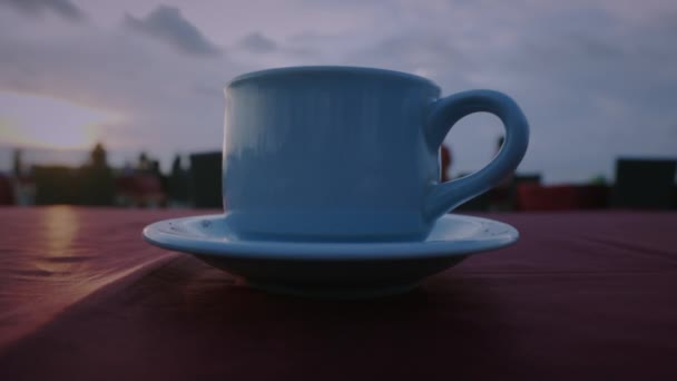 慢动作视频 在茶杯上观看咖啡或茶在桌子上在海滩 在日落时 在天空和大海的背景中 无法辨认的人们四处走动 — 图库视频影像
