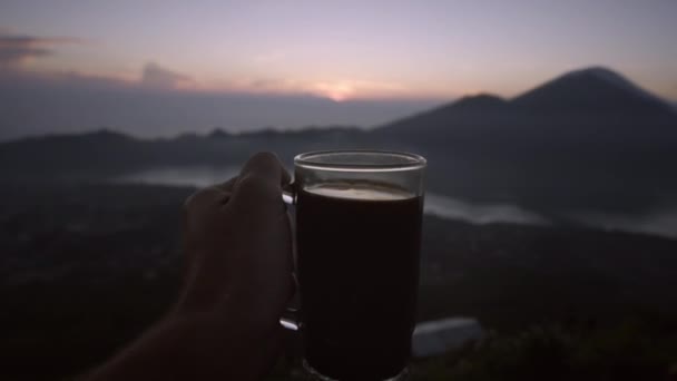 日の出を背景にコーヒーとガラスを手に持つ人の手 早朝のスカイラインと湖の上からの美しい眺め バリ島 インドネシア — ストック動画