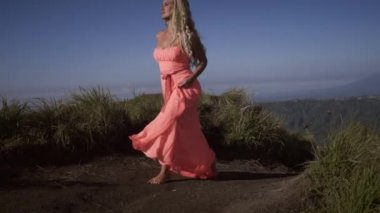 Siyah kum yolda yürüyen ve dağların, vadinin, gölün ve gökyüzünün arka planında volkanın tepesinde yükselen pembe elbiseli ciddi yalınayak genç kadının ön görünümü