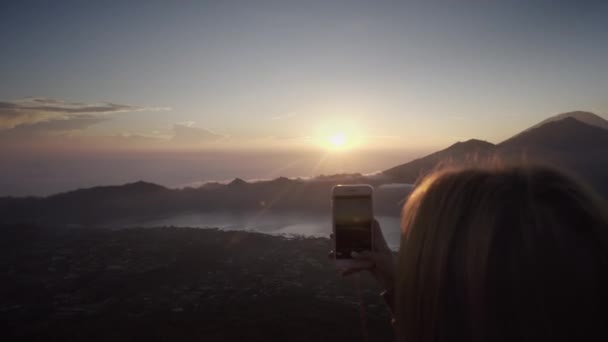 若い女性の手はスマートフォンを持ち 日の出を背景に写真を撮ります 早朝の湖や山の火山の頂上からの美しい眺め バリ島 インドネシア — ストック動画