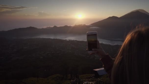 若い女性はスマートフォンを持ち 日の出を背景に写真を撮ります 早朝の湖や山の火山の頂上からの美しい眺め バリ島 インドネシア — ストック動画