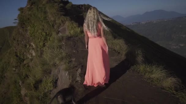 ピンクのドレスを着た裸足の若い女性のバックビューは 山を背景に火山の上に飼いならされた猿と黒い砂の道を歩きます — ストック動画