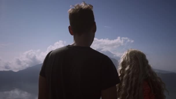 火山の頂上から素晴らしい景色を見ている男性とピンクのドレスを着た若い女性 端に立っている美しいカップルは 湖や空の背景に頭を回します — ストック動画