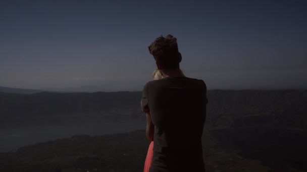 空を背景に山の端に立って抱きしめるカップル 火山の頂上から素晴らしい景色を見ている男性とピンクのドレスを着た若い女性 — ストック動画