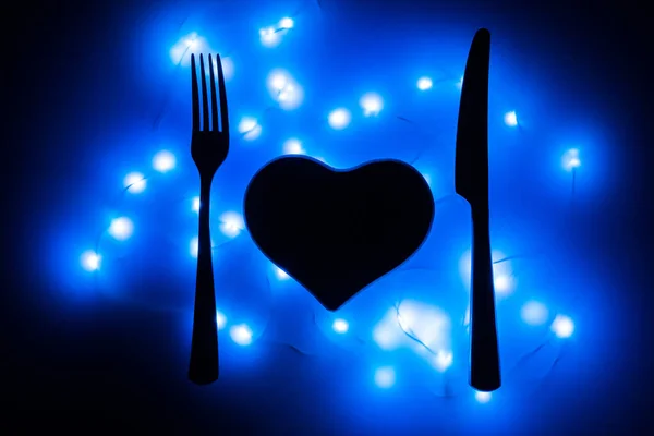 Black heart, fork and knife in the dark surrounded by blue garlands. The heart is served for dinner. Table setting for Valentine\'s day. Valentine\'s day February 14. Love is presented as food.