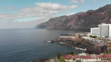 Hava atış. Dağ tatil beldesi birçok otel, palmiye ağaçları ve diğer bitki örtüsü. Mavi okyanus ve volkanik sahilde yenerek dalgalar. Bir yüksek katlı turist oteldir arka planda bir