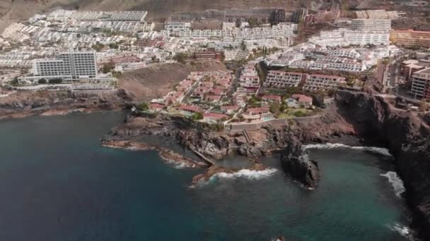 Disparo aéreo. Montanha resort cidade com muitos hotéis, palmeiras e outra vegetação. Oceano azul e as ondas batendo na costa vulcânica. Piscina natural em que os turistas não identificados nadar. Um enorme — Vídeo de Stock