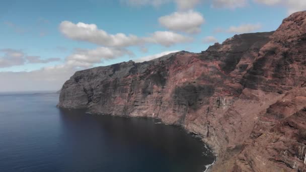 Disparo aéreo. A costa de uma enorme rocha vulcânica preta-vermelha é uma montanha nas águas azuis do oceano. Contra o céu azul. Ilhas Canárias, Tenerife, Los Gigantos — Vídeo de Stock