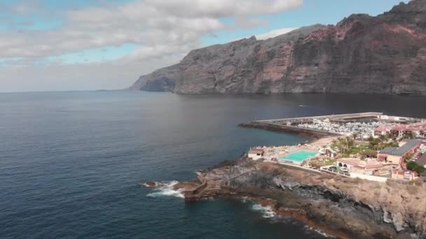 Disparo aéreo. Ciudad turística de montaña con muchos hoteles, palmeras y otra vegetación, al fondo un puerto deportivo y una enorme roca. Océano azul y las olas latiendo en la costa volcánica. Movimiento de cámara — Vídeo de stock