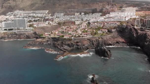 Disparo aéreo. Ciudad turística de montaña con muchos hoteles, palmeras y otra vegetación. Océano azul y las olas latiendo en la costa volcánica. Piscina natural en la que nadan turistas no identificados. Un enorme — Vídeo de stock