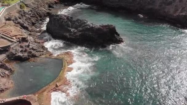 Disparo aéreo. Dia ensolarado sobre a costa do oceano e a costa vulcânica rochosa sobre a qual as ondas azuis estão batendo. Um enorme penhasco de rocha fica perto da costa. No contexto de uma piscina natural com — Vídeo de Stock