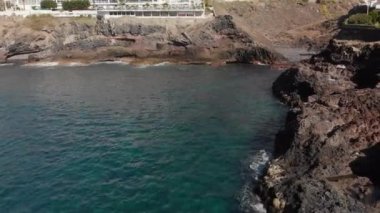 Hava atış. Uçuş geri volkanik ve kıyı. Manzara sarı ve kahverengi renk ve haklarında okyanus plaj turkuaz dalgalar dayak büyük taşlar var. Arka planda oteller şunlardır