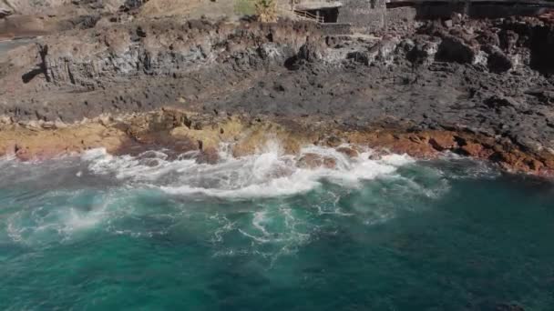 Luchtfoto schot. Leegstaande stenen gebouwen van oude mensen op de vulkanische kust van het eiland. Het landschap is grote stenen van gele en bruine kleur en turquoise golven slaan over hen op het strand — Stockvideo