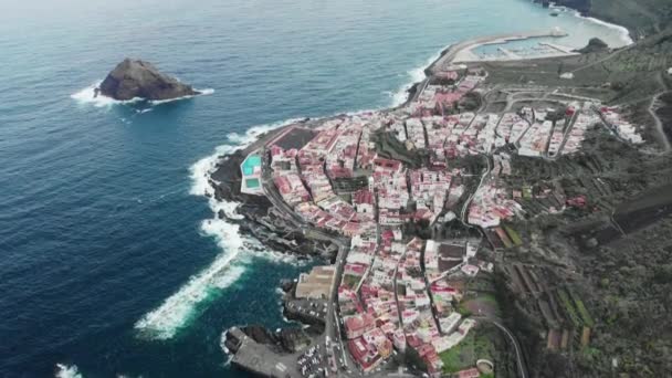 Un disparo aéreo. Larga costa, fuertes olas. Océano azul y costa volcánica. Un pequeño pueblo con casas blancas y techos rojos a orillas del Océano Atlántico, cerca de la base de las montañas. Una piedra enorme — Vídeos de Stock