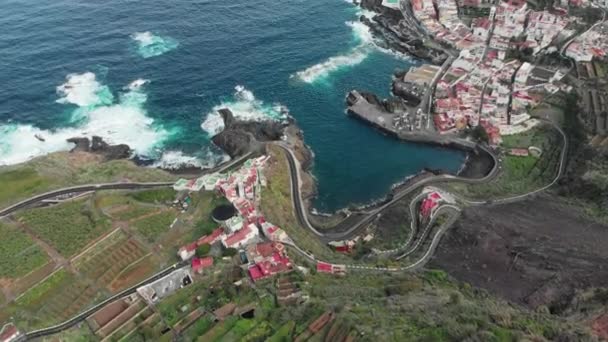 Aerial shot. Small town at the base of the mountain, ocean and volcanic coastline. Cars driving on mountain winding roads serpentine hairpins. The concept of vacation and vacation on the ocean — Stock Video