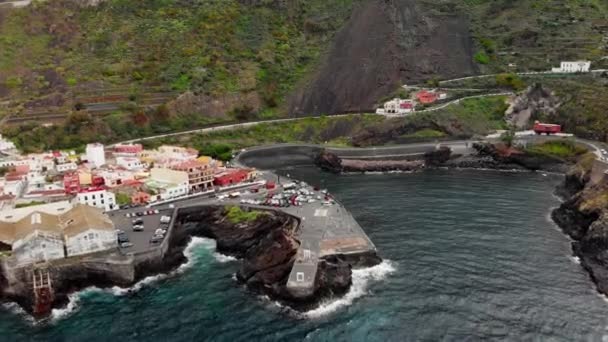 Légi felvétel. Kis település a hegyek és a vulkán tövében. Vulkanikus köves óceán partján, fehér hab dúl, feltűnő a tengerparti reef hullámok. Garachico, Tenerife, Kanári-szigetek — Stock videók