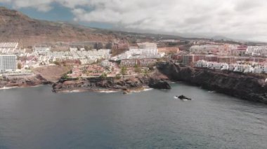 Hava atış. Dağ tatil beldesi birçok otel, palmiye ağaçları ve diğer bitki örtüsü. Mavi okyanus ve volkanik sahilde yenerek dalgalar. büyük taş ön planda. Tepe üstündeki yanardağ