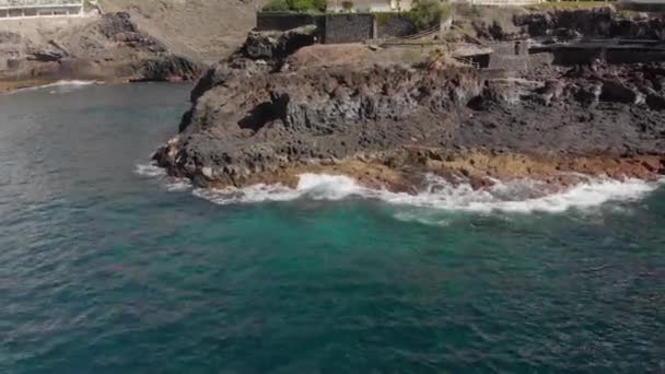 Aerial shot. Approaching the volcanic coast on which two unrecognized tourists take pictures. The landscape is big stones of yellow and brown color and beating about them turquoise waves on the beach — Stock Video