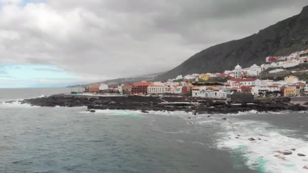 Aerial shot. Volcanic stone coast of frozen lava against which hit the blue ocean waves with white foam. Small town at the base of the mountain. Cars driving on the road near the sea. Garachico — Stock Video