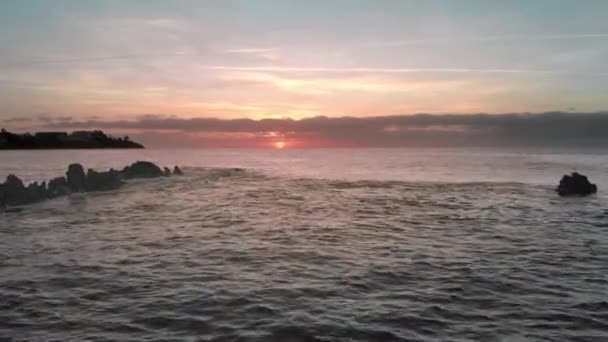 Κεραία βολή. Ανατολή του ηλίου πάνω από τον ωκεανό. Ροζ και πορτοκαλί χρώματα. Νησί του ωκεανού, κύματα βουτιά σε ηφαιστειακές πέτρες λάβας κατεψυγμένα που προεξέχουν από το νερό. Στο φόντο είναι αρκετά αγνώριστος — Αρχείο Βίντεο