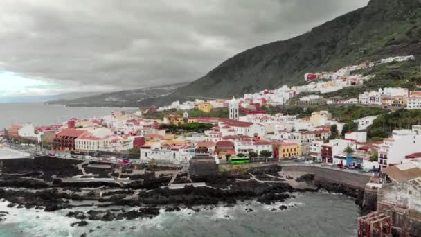 Un disparo aéreo. Pequeño pueblo en la base de las montañas y el volcán. Orilla volcánica del océano rocoso, espuma blanca que golpea las olas en el arrecife costero. Garachico, Tenerife, Islas Canarias. la cámara se va — Vídeo de stock