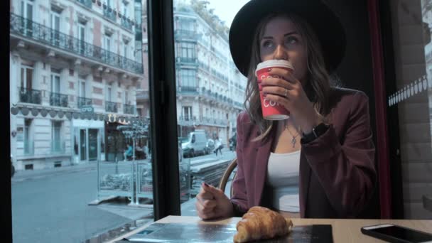 Una bella donna d'affari francese con un cappello sta bevendo caffè in una caffetteria sullo sfondo di una strada francese. Sul tavolo c'è un croissant. Rallentatore — Video Stock