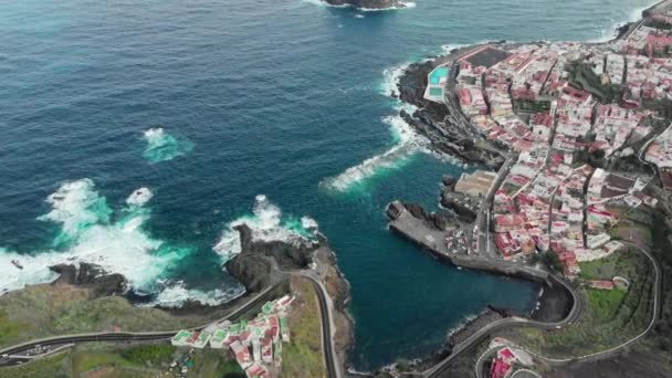 Tiro aéreo. Pequena cidade na base da montanha, oceano azul e costa vulcânica. Carros dirigindo em estradas sinuosas montanha serpentina grampos de cabelo. Piscinas naturais, nuvens de baixo volume, pedra vulcânica em — Vídeo de Stock