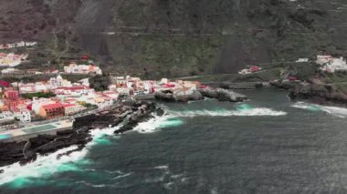 Hava atış. Hangi karşı donmuş lav volkanik taş sahil mavi okyanus dalgaları beyaz köpük ile vurmak. Beyaz evler ve dağın dibinde bir tapınak ile küçük bir kasaba. Üzerinde sürüş araba
