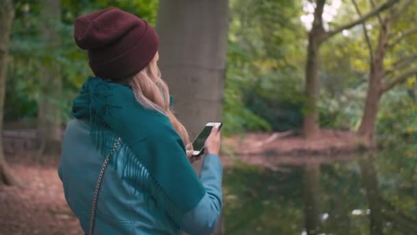 Медленное движение Девушка в красной шляпе и синей куртке, осенью в парке у озера вытаскивает мобильный смартфон и фотографирует красивый пейзаж — стоковое видео