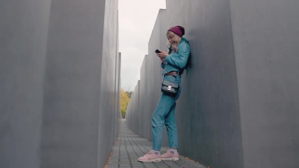 スローモーション白人少女観光は、コンクリートのような近代的な都市の建物の巨大な灰色のスラブ clawing 立っています。笑顔し、モバイルのスマート フォンを使用します。孤独との損失の概念 — ストック動画