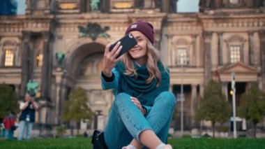 Sonbaharda Kafkas turist kızı Berlin, Almanya Katedrali yakınlarında bir çim üzerinde oturan akıllı telefon üzerinde selfie fotoğraf yapar. Yavaş hareket
