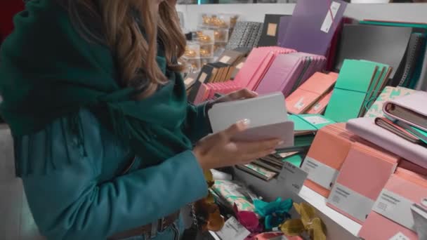 Hermosa chica caucásica en una chaqueta azul y una bufanda verde, elige y compra suministros de oficina y cuadernos en la tienda, cámara lenta — Vídeos de Stock