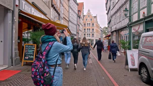 Hannover, Németország-október 2018: turista lány egy hátizsák áll a közepén egy keskeny európai utca és fényképeket a smartphone. Az emberek elhaladnak a lány. Az Európai Unió történelmi központja — Stock videók