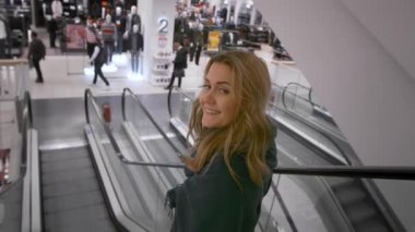 Güzel beyaz kız gülümsüyor, alışveriş merkezinde yürüyen merdivenden aşağı gidiyor. Tanınmayan alıcıların arka planına karşı. Yavaş çekim