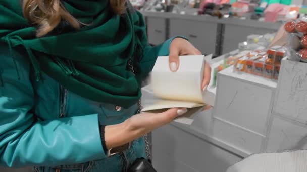 Hermosa chica caucásica en una chaqueta azul y una bufanda verde, pasa las páginas de un cuaderno en una tienda de papelería, cámara lenta. El concepto de película animada y dibujos animados — Vídeo de stock