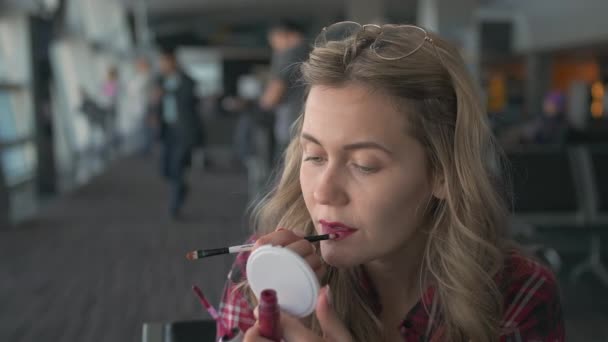 Slow Motion Una joven hermosa con una camisa a cuadros roja, sentada de perfil, se aplica lápiz labial en los labios con un cepillo y se mira a sí misma en un pequeño espejo sentado en la zona de salida de la — Vídeo de stock