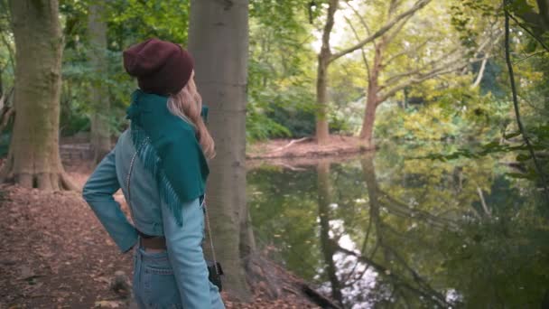 Yavaş Hareket Kırmızı şapkalı ve mavi ceketli genç bir kadın kız, göl e yakınlarındaki bir parkta sonbaharda, kameraya sırtını döner ve güzel manzaraya bakarak doğaya hayran kalır. — Stok video