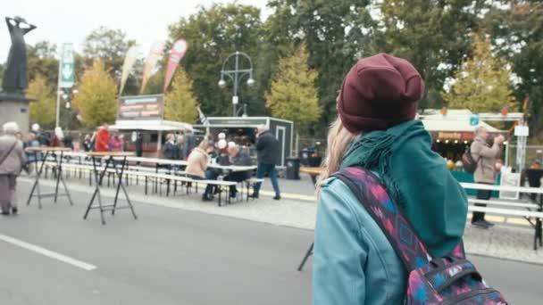 Yavaş Hareket Mavi ceket ve arkasında bir sırt çantası ile Genç Kafkas kız turist şehir fuarı etrafında yürüyor. Tanınmayan dükkan ve insanların arka planına karşı — Stok video