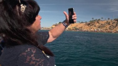 Tropik bir ada yakınlarındaki bir teknede Kıdemli kadın yetişkin turist video iletişim konuşmak için bir akıllı telefon kullanır. Bir üst yaş blogger kavramı. Yavaş Çekim