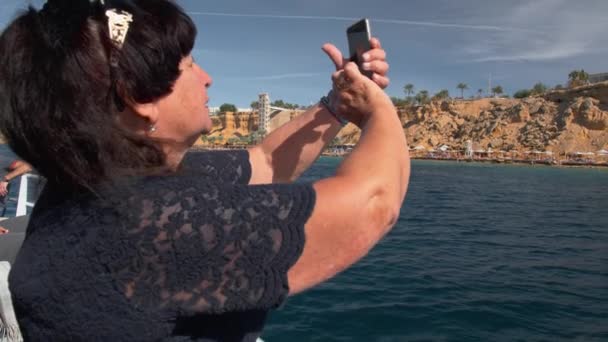 Senior mujer feliz turista adulto en un barco utiliza un teléfono inteligente móvil para obtener fotos de una isla tropical. Moción lenta — Vídeos de Stock