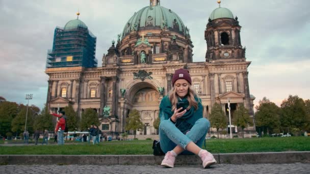 Berlín, Německo-říjen 2018: turistická dívka na podzim používá mobilní telefon a současně sedí na trávníku nedaleko katedrály v německém Berlíně. Zpomaleně — Stock video