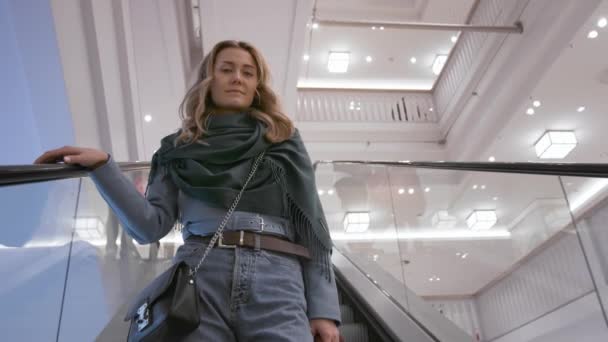 Hermosa chica caucásica en una chaqueta azul y una bufanda verde, sonriendo, bajando por la escalera mecánica en un centro comercial. Vista inferior, cámara lenta — Vídeos de Stock