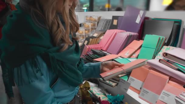 BERLÍN, ALEMANIA - Octubre 2018: Hermosa chica caucásica con una chaqueta azul y una bufanda verde, elige y compra bloc de notas en la papelería, cámara lenta — Vídeos de Stock