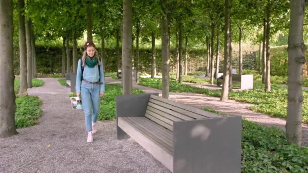 Hannover, Alemanha. Uma jovem turista com um mapa nas mãos e uma mochila sentada em um banco de pedra no parque. Abre um mapa em papel e olha o caminho nele. Na armação um monte de árvores. Movimento lento — Vídeo de Stock