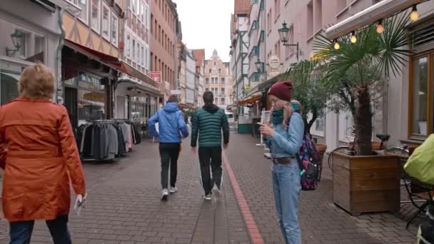 HANOVER, ALEMANIA - Octubre de 2018: una turista con una mochila está parada en medio de una estrecha calle europea y utiliza un smartphone. La gente pasa. El concepto de vida virtual. El histórico — Vídeo de stock