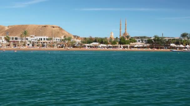 Пляж і туристи на курорті біля прекрасного моря влітку в сонячний день. На тлі пальм і готелів. На відстані видно мінарет і мечеть. Повільний рух — стокове відео